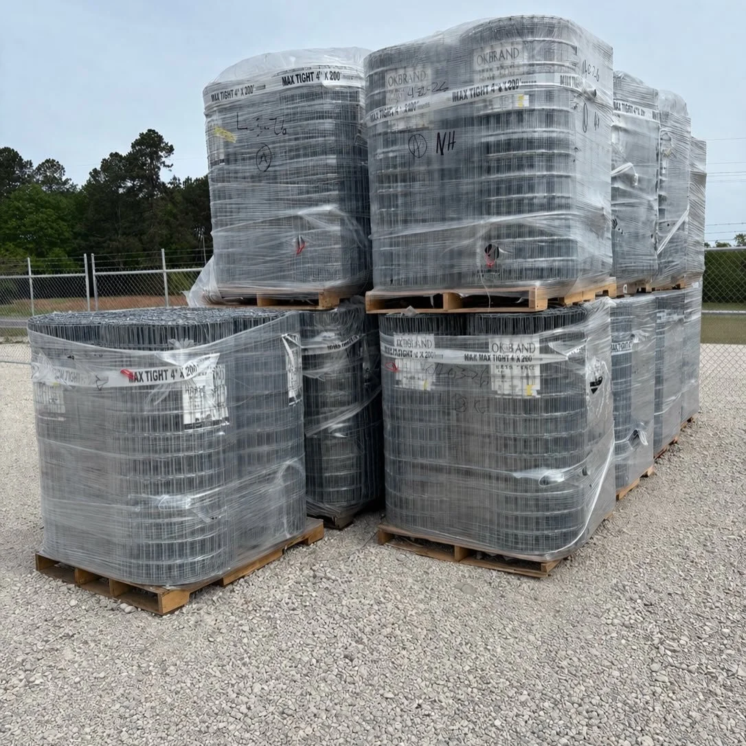Stacked pallets of coiled wire fencing wrapped in plastic, placed on a gravel lot next to a chain-link fence with trees in the background.