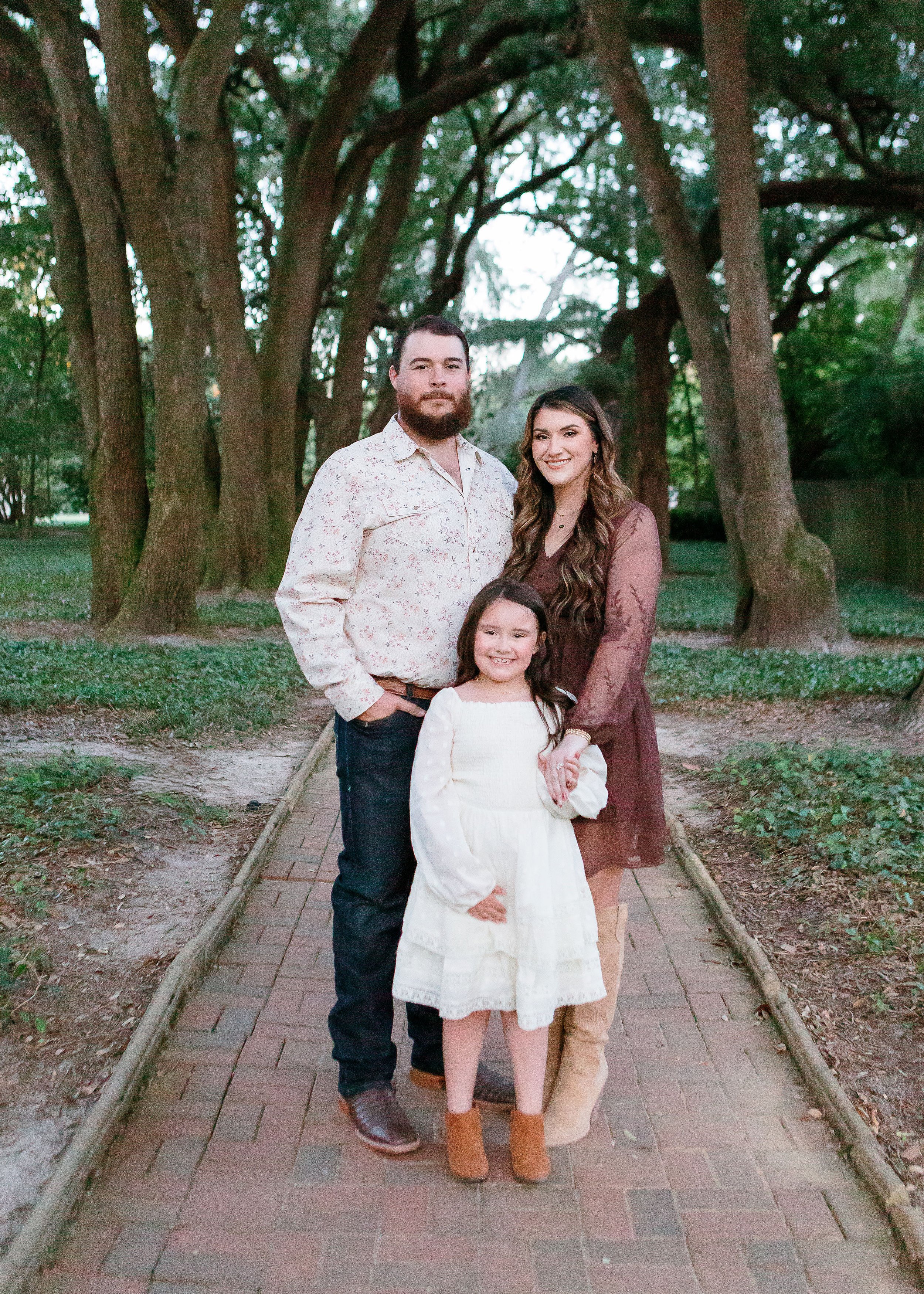 A family of three posing on a brick path in a park with tall trees in the background. The man is standing on the left, wearing a floral shirt and dark jeans. The woman is on the right, wearing a brown dress and tan boots, with her arm around the young girl in front. The girl is smiling, wearing a white dress and tan boots.