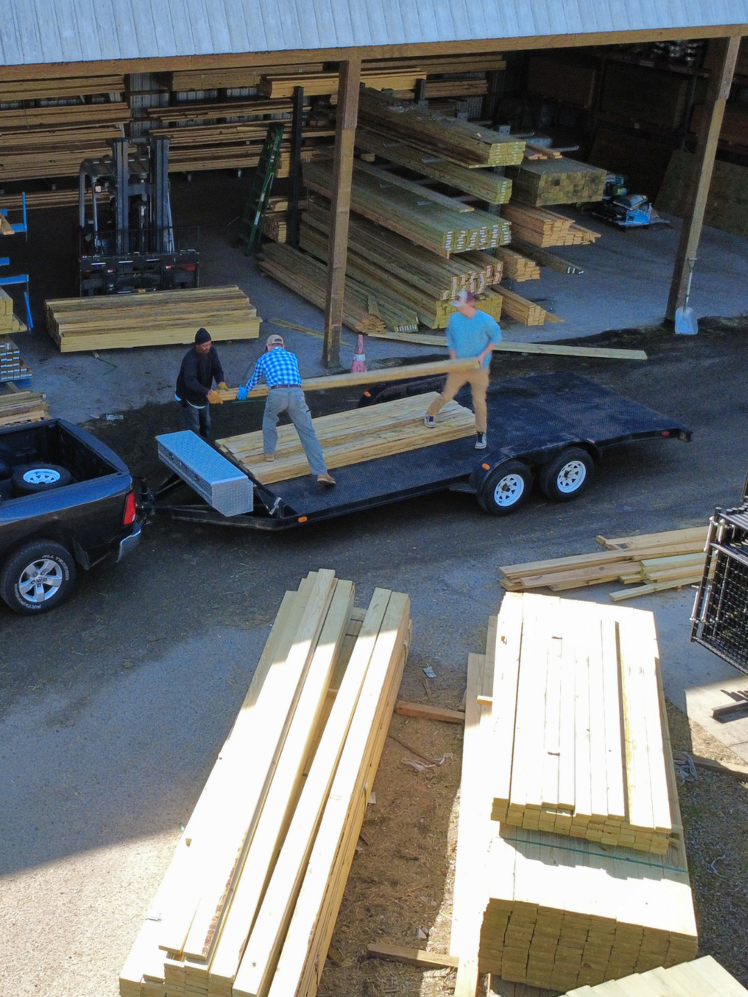 Workers lifting a wooden plank onto a trailer at a lumber yard, with stacks of lumber and construction materials in the background.