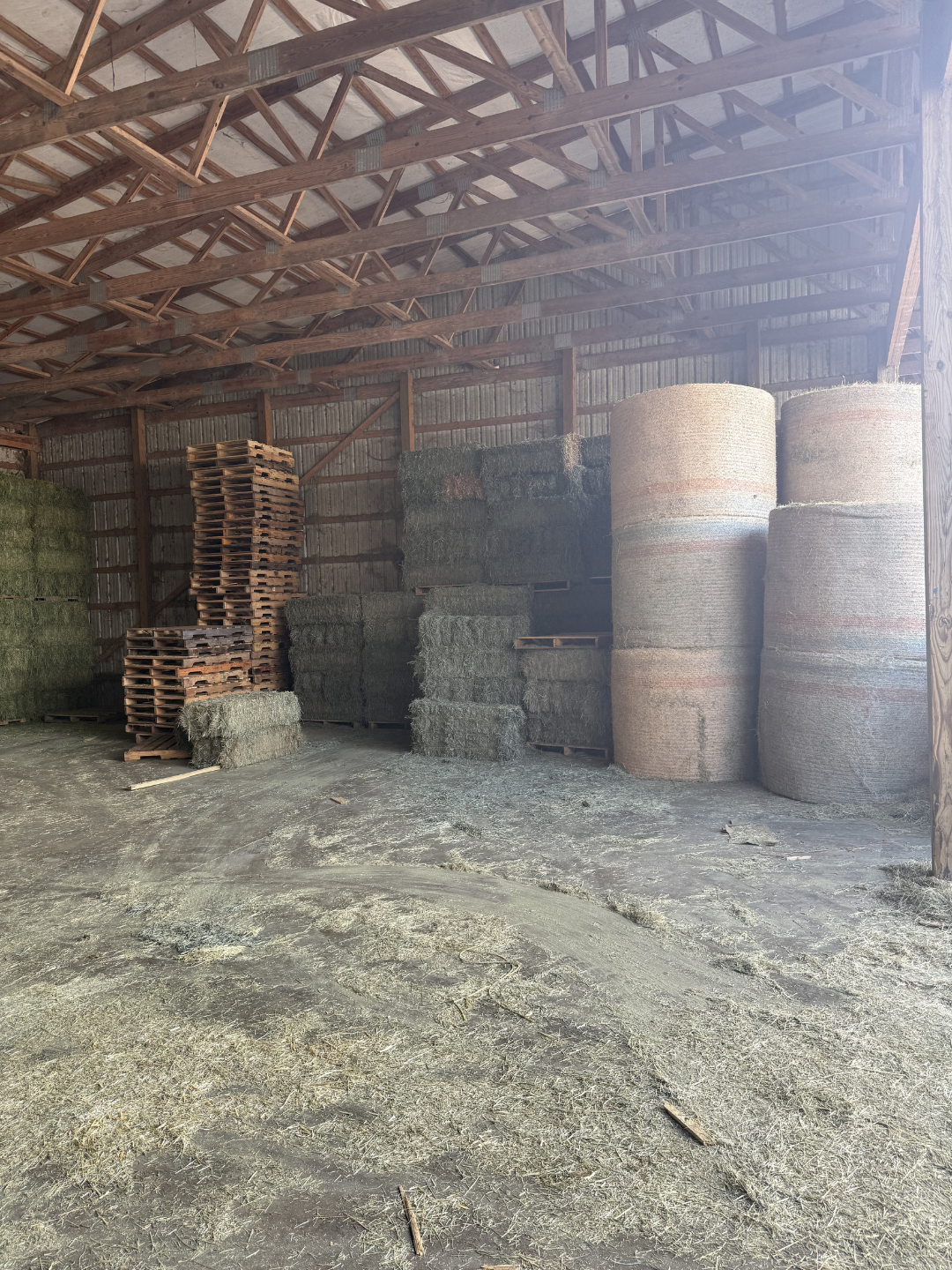 Stacks of hay bales and wooden pallets inside a barn with wooden beams and a dirt floor.