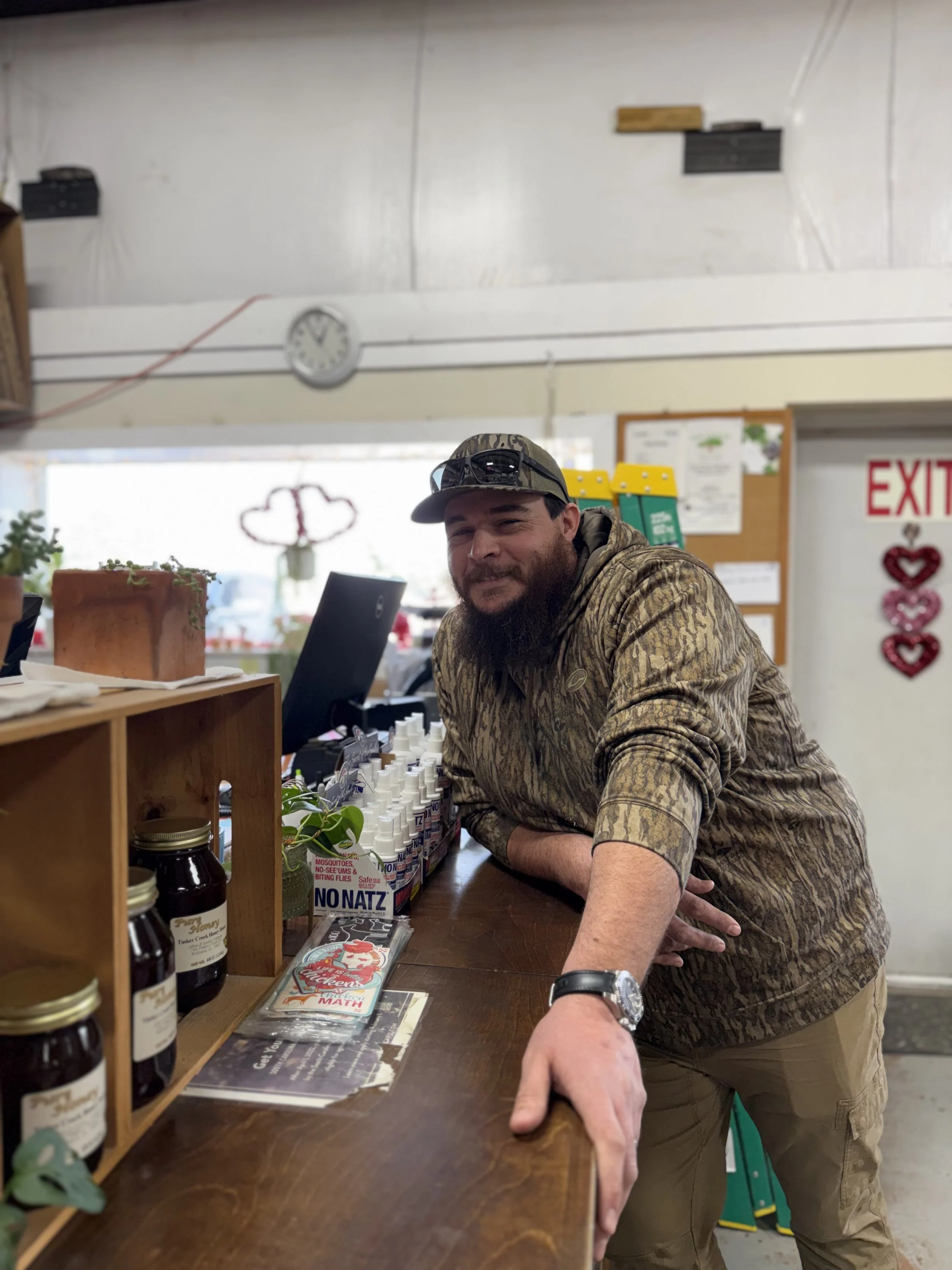 A bearded man in camouflage-patterned hoodie leaning on a wooden counter in a store, smiling at the camera. Behind him are jars and bottles, with a computer monitor and small plants nearby. In the background, there is a window and wall with a clock, colorful decorations, and an exit sign.
