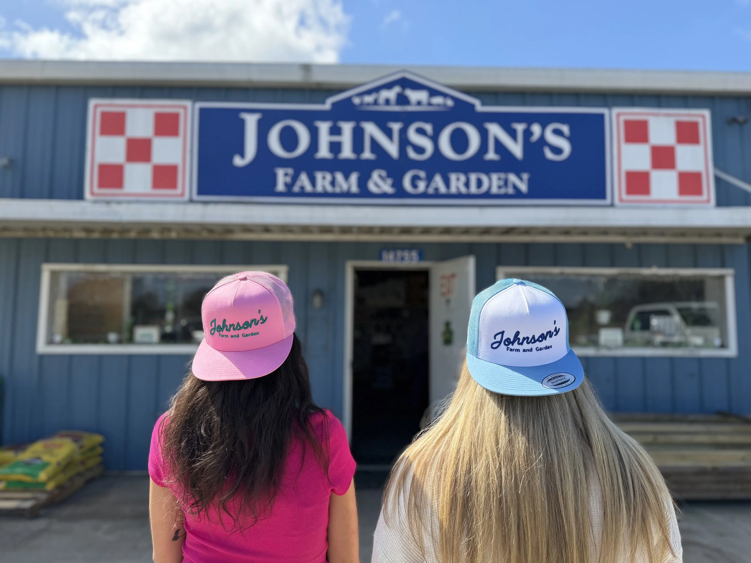 Two women are standing in front of Johnson's Farm & Garden store, facing away. One wears a pink hat and shirt, the other a blue and white hat. The store's sign is visible above them.