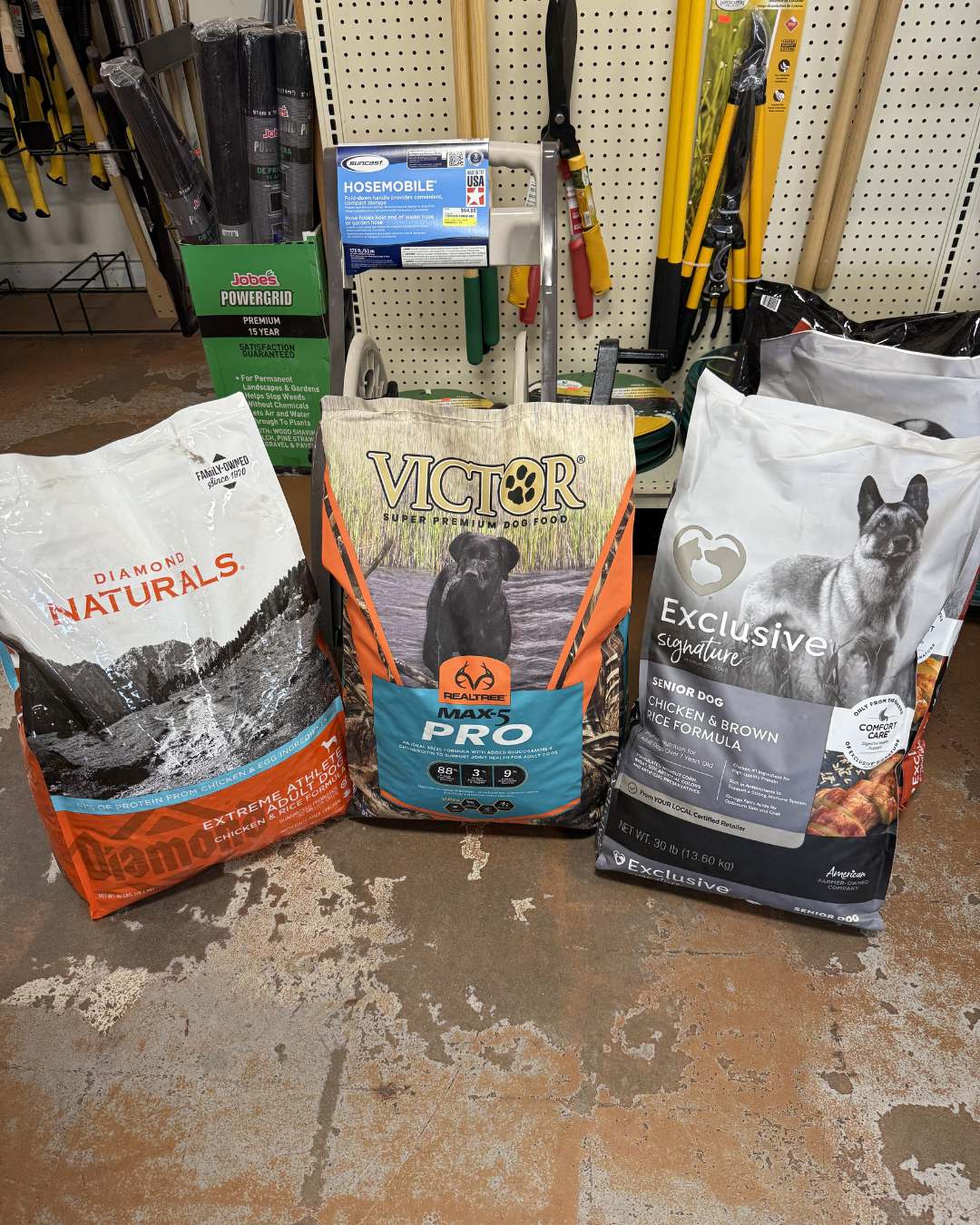 Three large bags of dog food placed on a concrete floor in a hardware store. The left bag is Diamond Naturals with a mountain landscape, the middle bag is Victor with a picture of a black dog, and the right bag is Exclusive Signature featuring a Belg