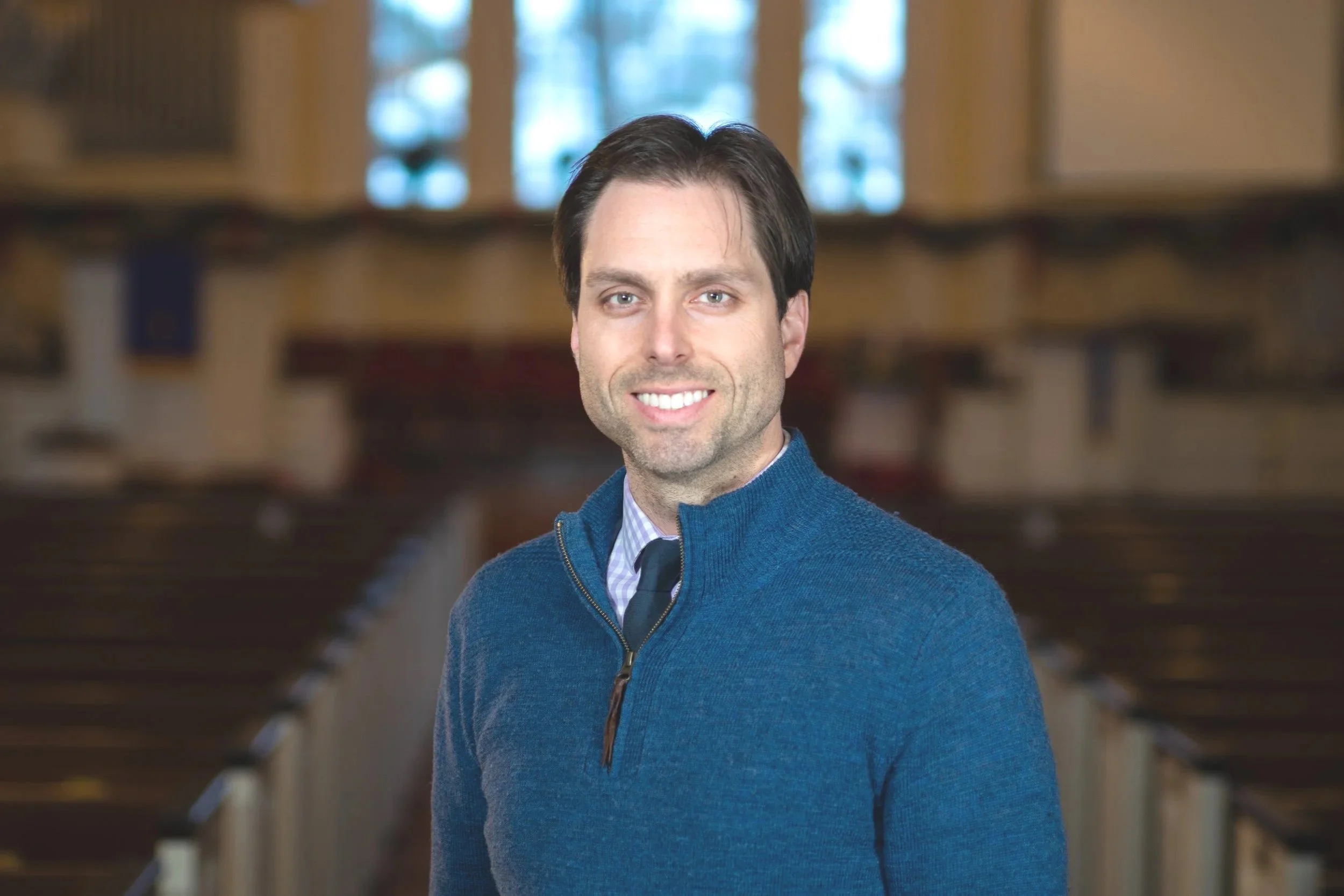 A man with dark hair and blue eyes, wearing a blue zip-up sweater over a collared shirt and tie, standing inside a church or chapel with wooden pews and stained glass windows in the background.