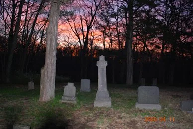 Sunset over a graveyard with headstones and a tall monument among leafless trees