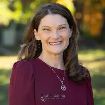 A woman with brown hair smiling outdoors, wearing a maroon blouse and a peace sign necklace.