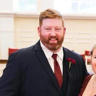 A smiling man with red hair and beard wearing a black suit, white shirt, and red tie, standing indoors with a blurred background.