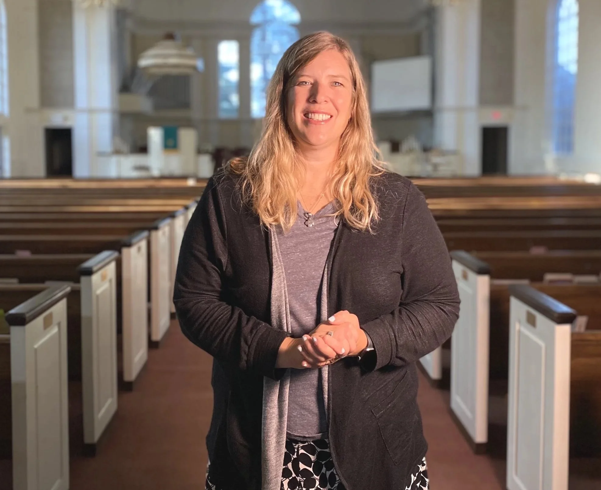 Woman smiling standing inside a church with wooden pews and large arched windows.