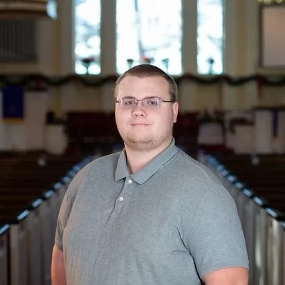A man with glasses and short hair stands inside a church or auditorium, facing the camera with a neutral expression.