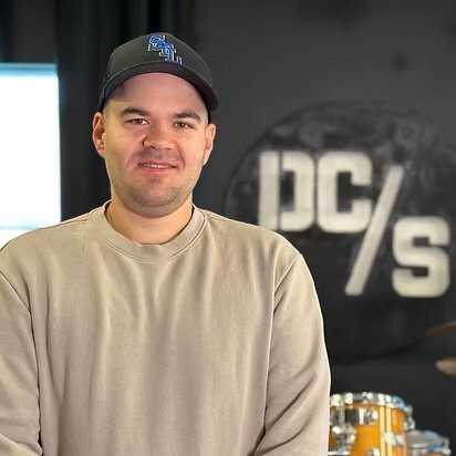 A young man wearing a black baseball cap and beige sweatshirt, standing in front of a black wall with a large, partially visible 'DC//' sign.