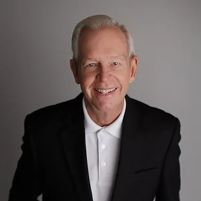 Portrait of a smiling middle-aged man with gray hair, wearing a black blazer and white shirt, against a gray background.