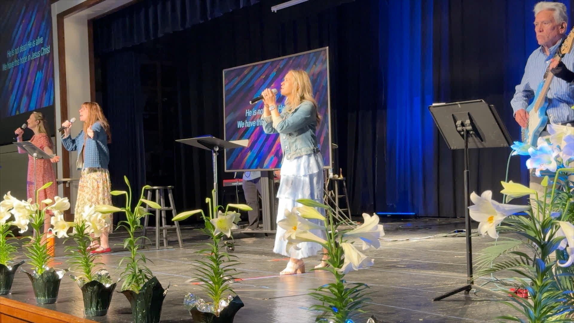 People singing on stage during a church service with floral decorations and lyrics displayed on a screen.