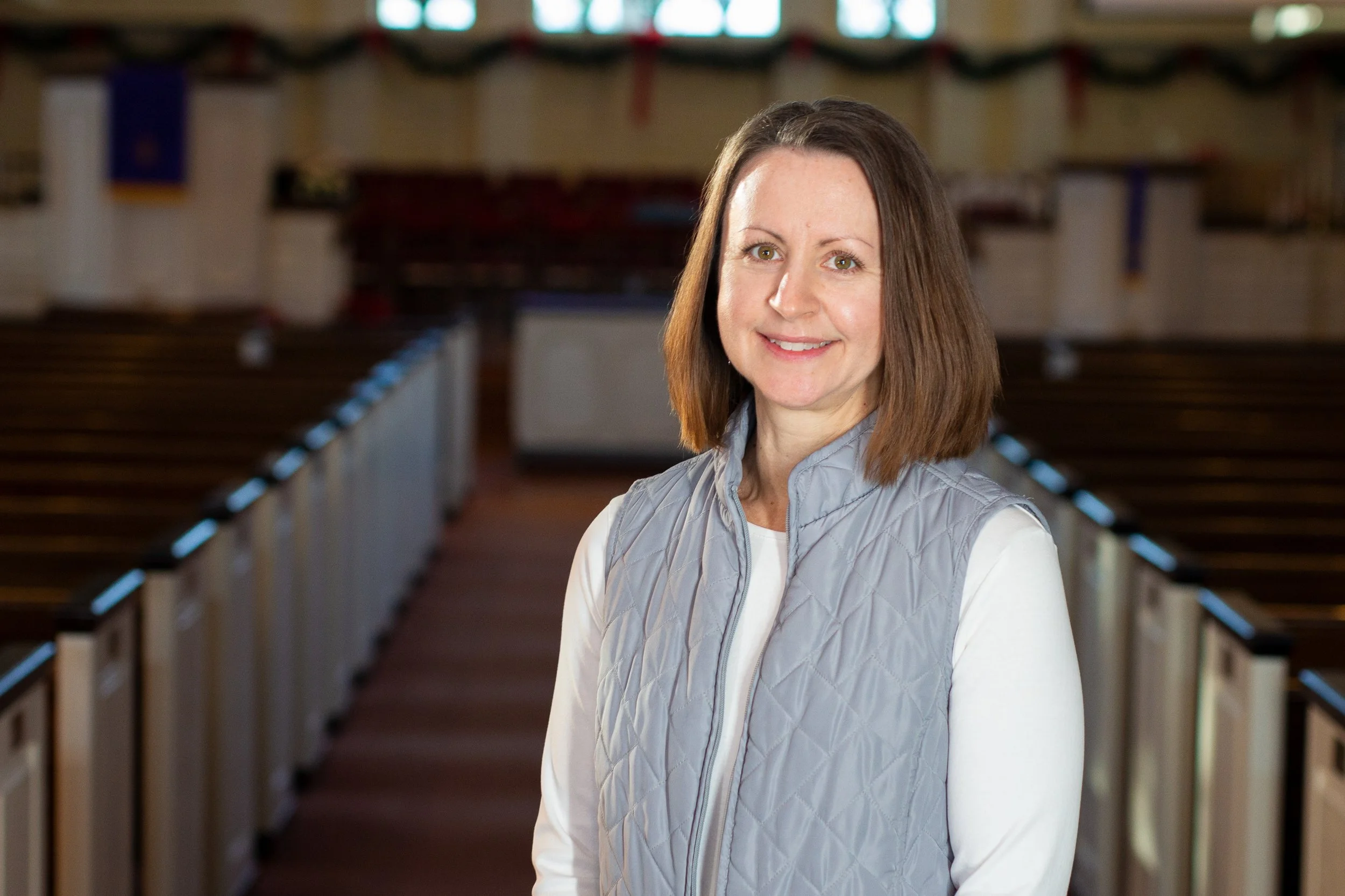 A woman with shoulder-length brown hair, wearing a light gray quilted vest over a white shirt, standing in an indoor arena with empty wooden pews and American flags in the background.