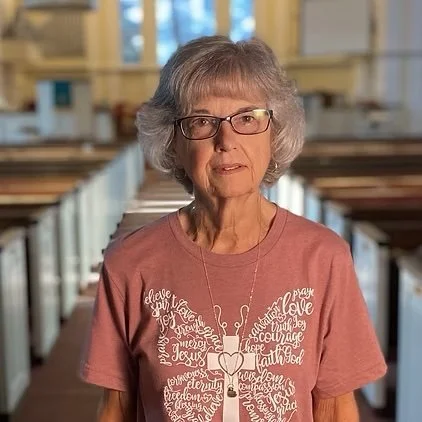 An elderly woman with glasses and gray hair standing indoors, wearing a pink T-shirt with a heart-shaped tree graphic and religious words, in a spacious room with wooden structures.