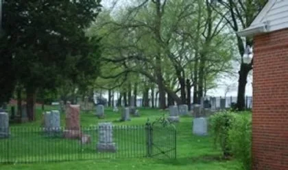 Graveyard with headstones surrounded by green grass and trees, partially enclosed by a black iron fence.