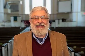 An elderly man with glasses and a beard, smiling, in an indoor setting with chairs and windows in the background.