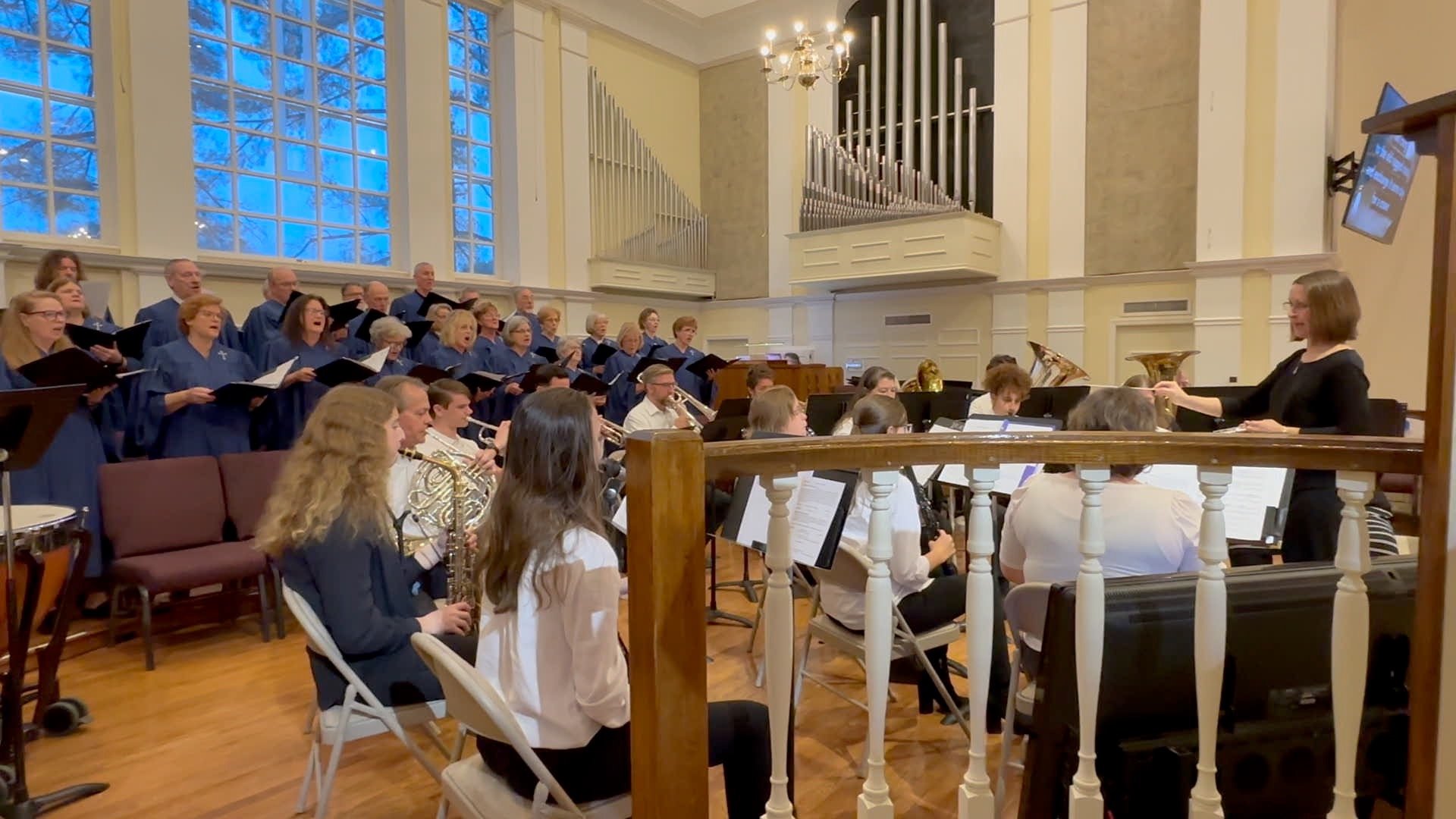 Chorus and orchestra performing in a church or concert hall with large windows and organ pipes in the background, directed by a woman conductor.