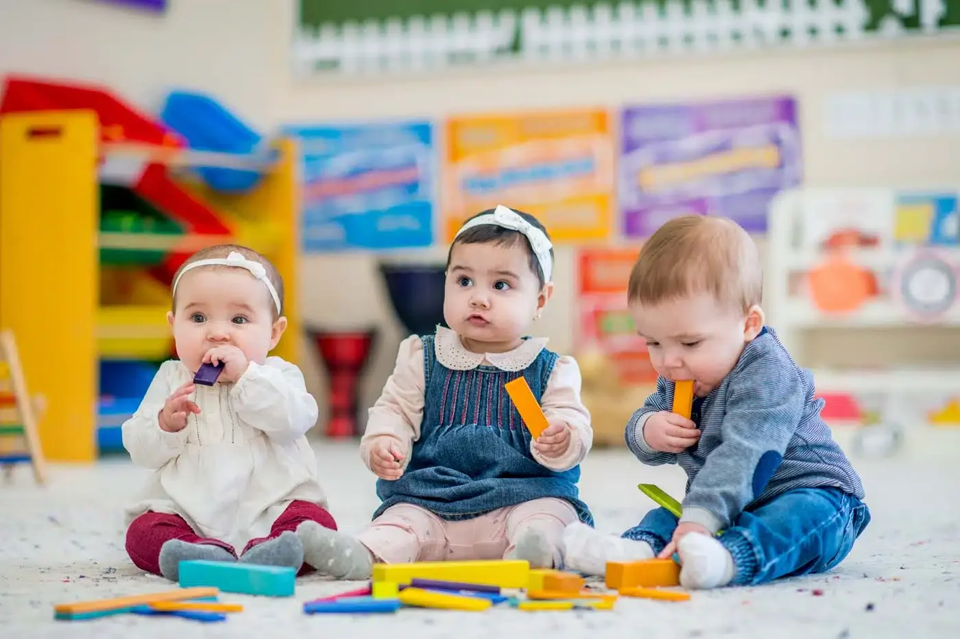 Three babies sitting on the floor in a colorful playroom, playing with building blocks. The background features shelves with educational toys and posters.
