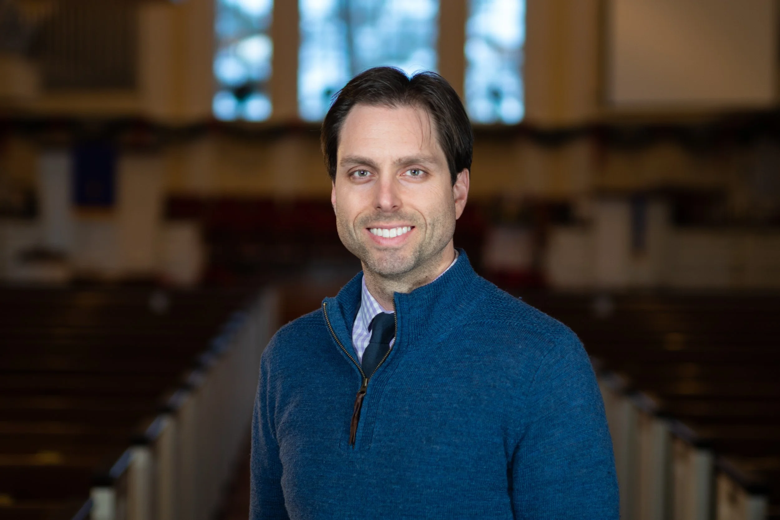 A man with dark hair and blue eyes, wearing a blue zip-up sweater over a collared shirt and tie, standing inside a church or chapel with wooden pews and stained glass windows in the background.