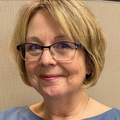 A middle-aged woman with blonde hair, glasses, and a light blue top smiling at the camera.