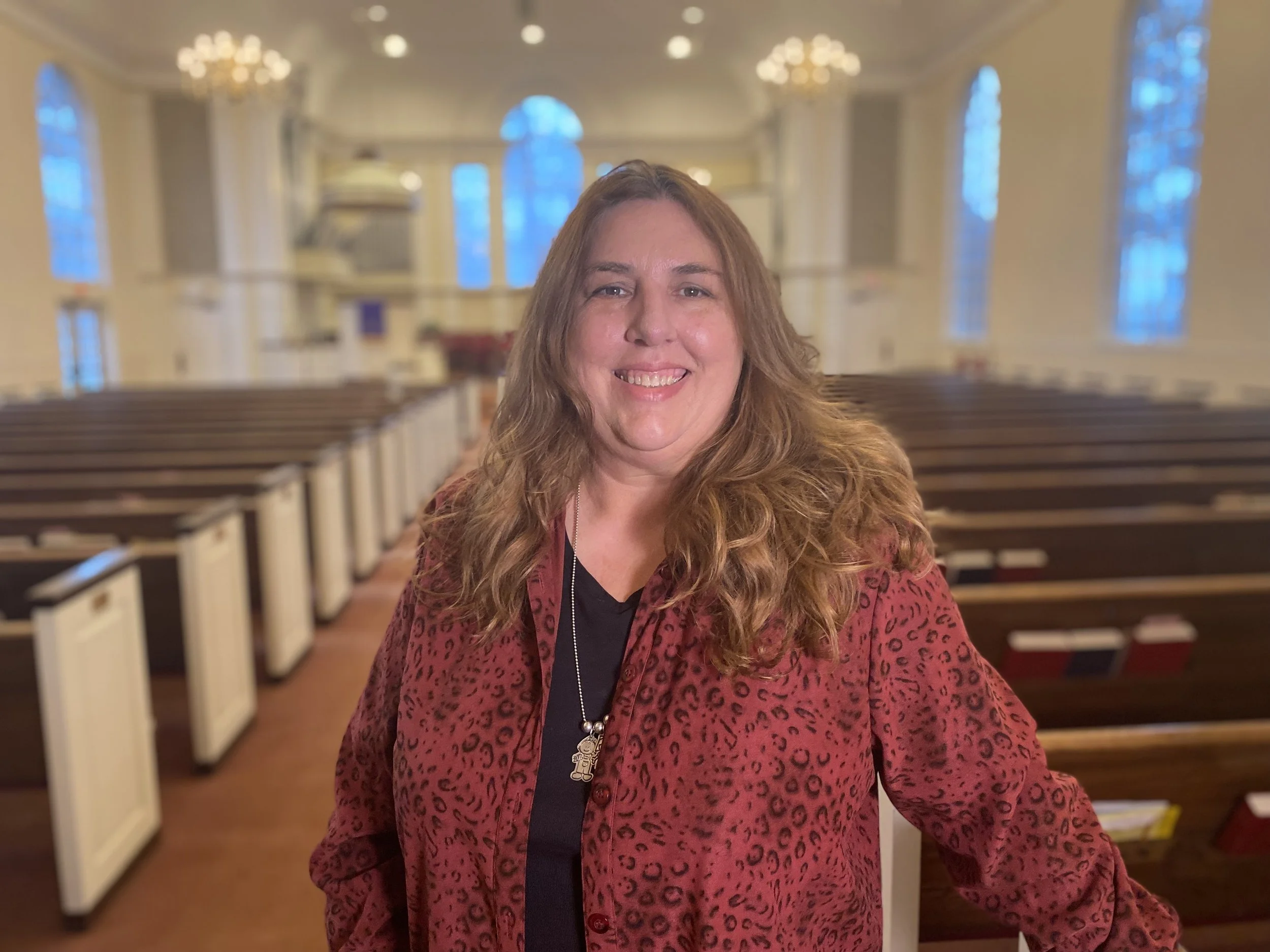A woman with long wavy brown hair smiling inside a church with high ceilings, multiple large arched stained glass windows, chandeliers, and wooden pews.