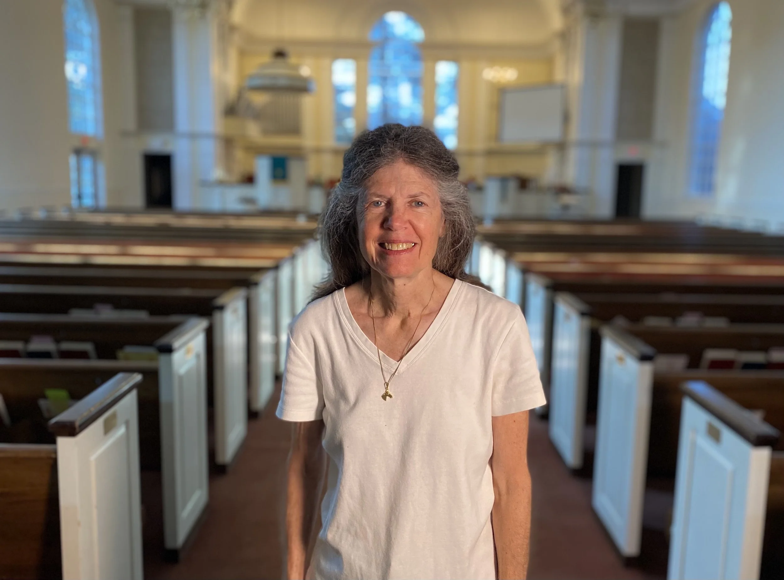An elderly woman with gray hair, sitting in a church pew, smiling, wearing a white shirt and a necklace with a small pendant.