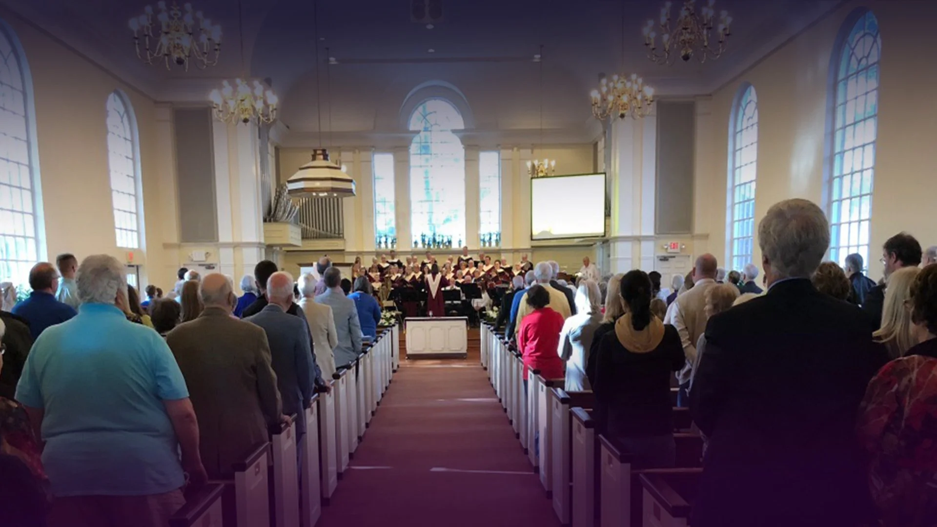 A congregation gathered inside a church with tall arched windows, chandeliers, and a choir at the front during a service or ceremony.