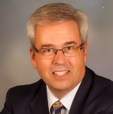 A middle-aged man with gray hair, glasses, and a friendly smile wearing a dark suit, white shirt, and a patterned tie.