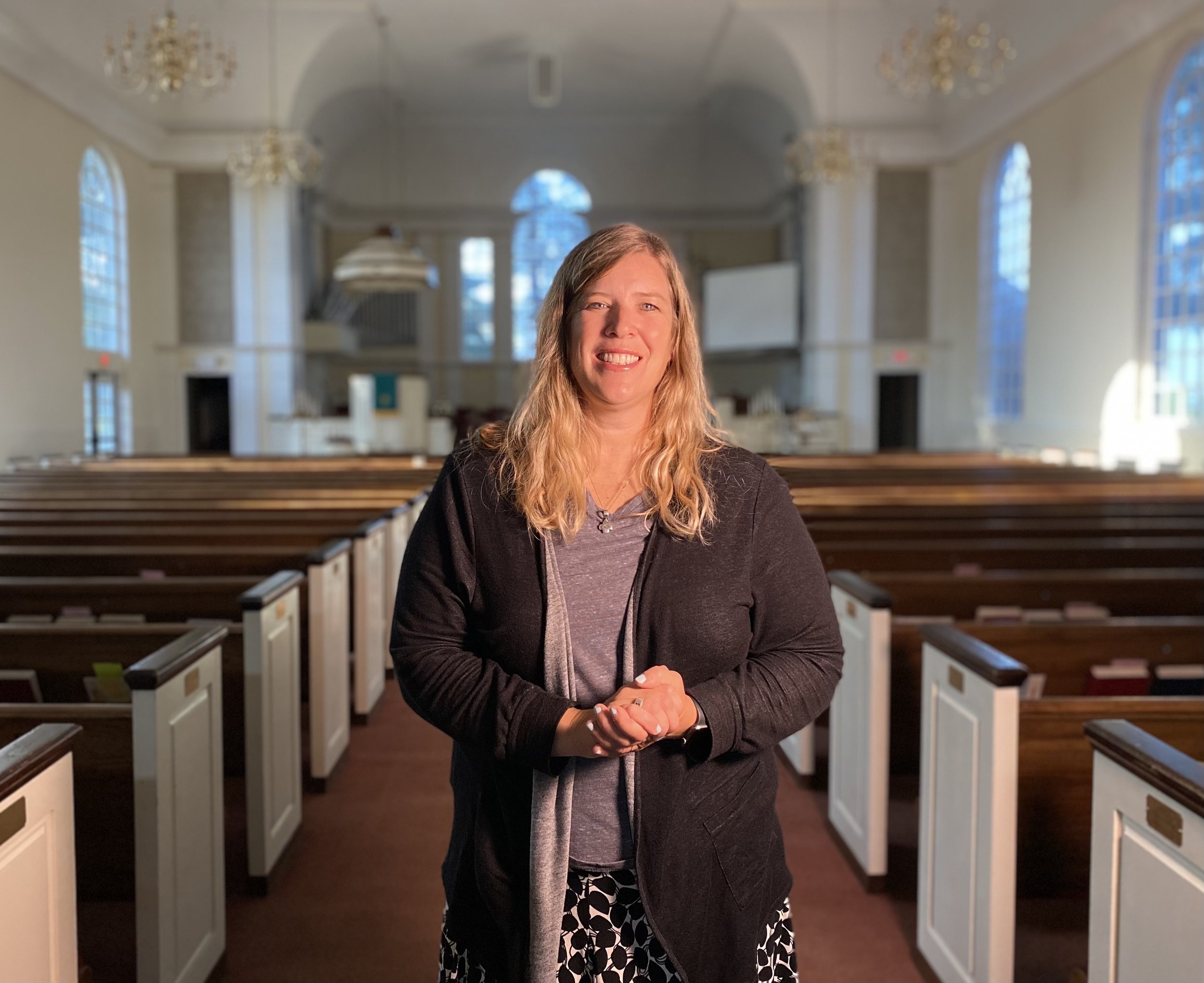 Woman smiling standing inside a church with wooden pews and large arched windows.