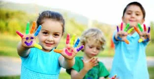 Three children outdoors showing their painted hands with colorful finger paint, smiling and having fun.