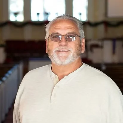 An older man with gray hair and beard, wearing glasses and a light-colored shirt, standing indoors with blurred windows behind him.
