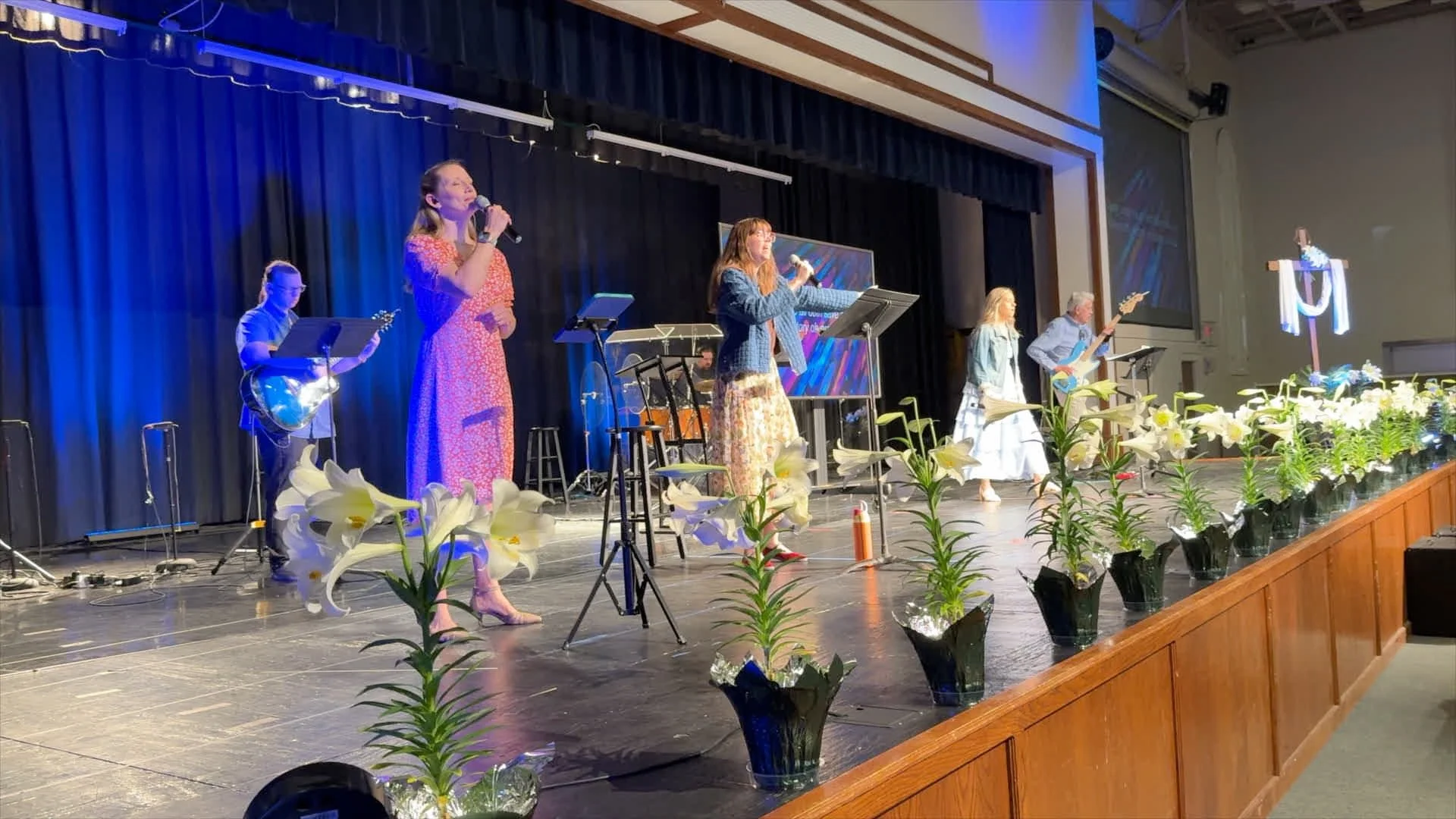A group of five musicians performing on a stage decorated with white flowers, including lilies, during a concert with a blue and black curtain background. There are women singing into microphones, a guitarist, and other band members, with music stands and equipment visible.