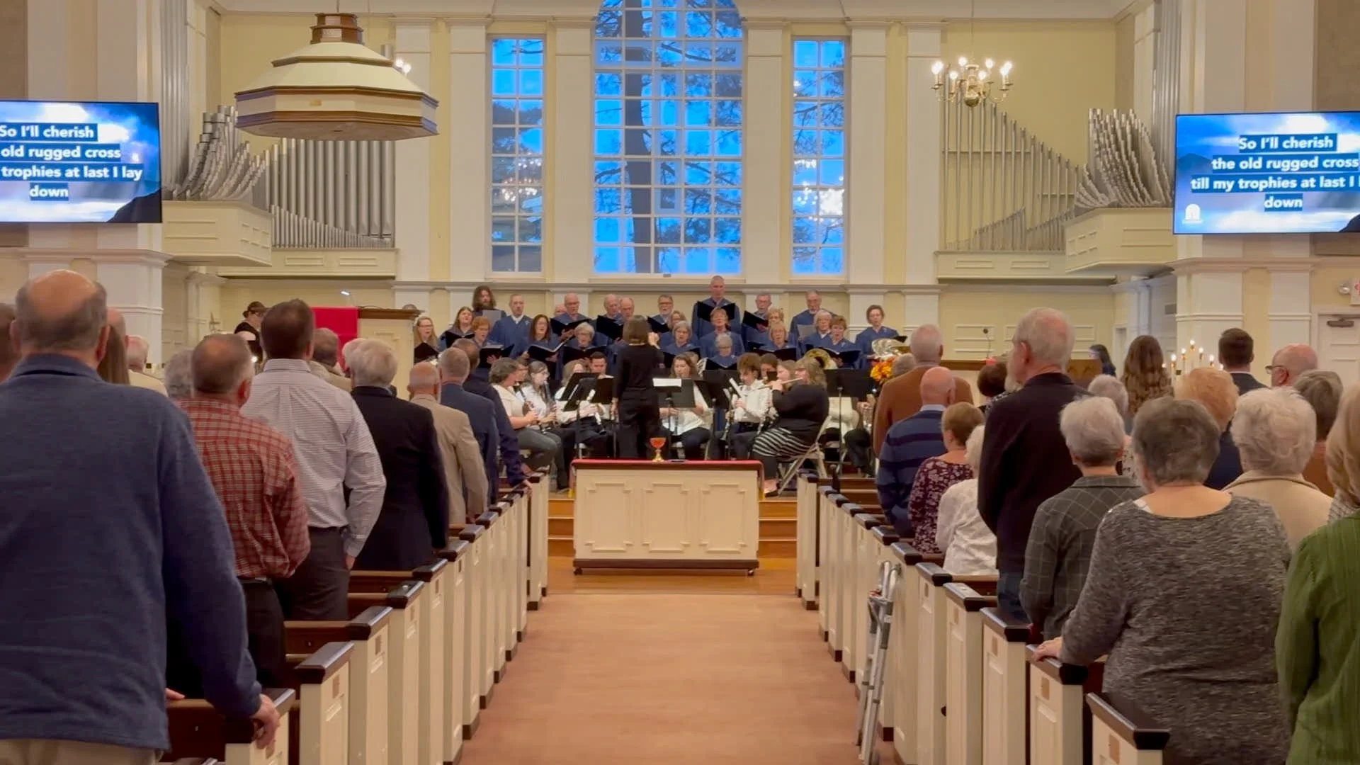 A choir and orchestra perform in a church with a large audience watching. The church has tall windows, chandelier lighting, and music screens displaying lyrics.