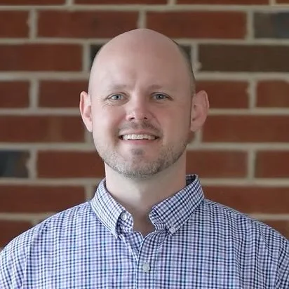 Portrait of a bald man with a beard, smiling, wearing a checkered shirt, standing in front of a brick wall.