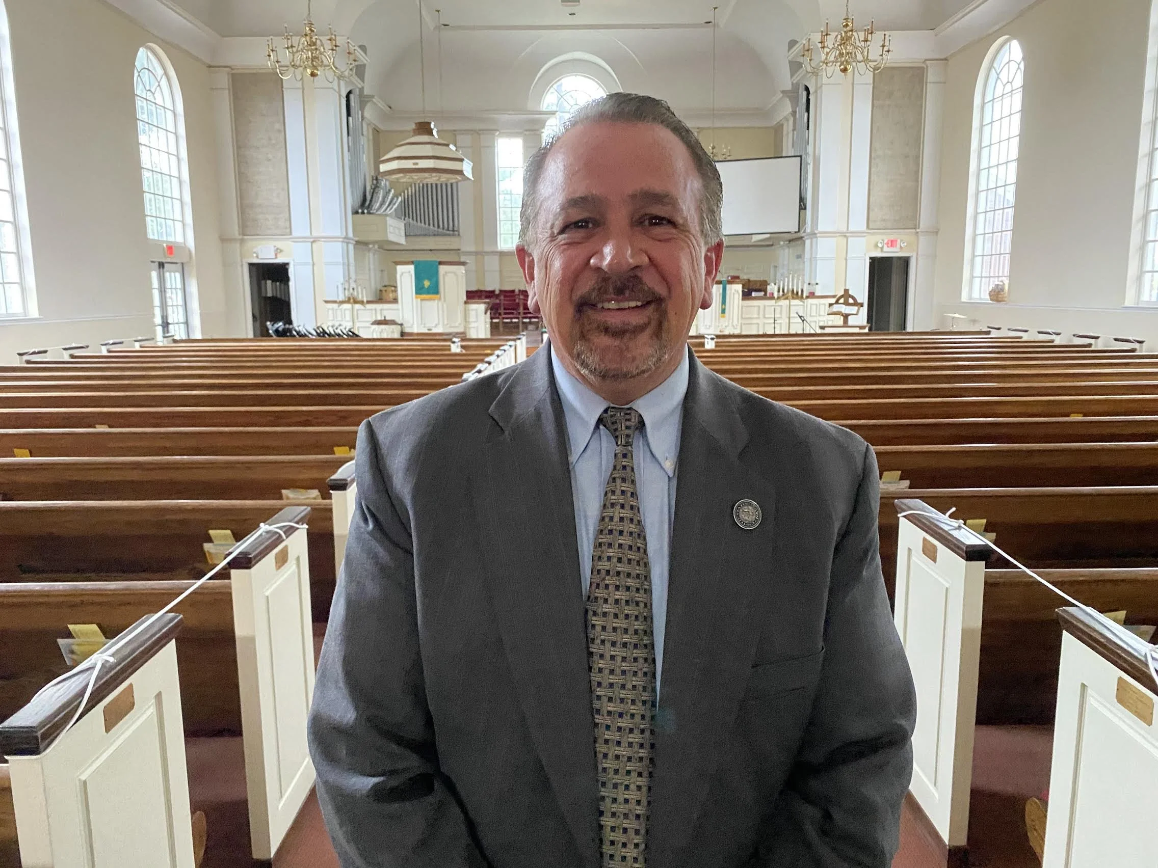 Smiling man in a gray suit standing inside a church with empty wooden pews, large arched windows, chandeliers, and a church altar in the background.