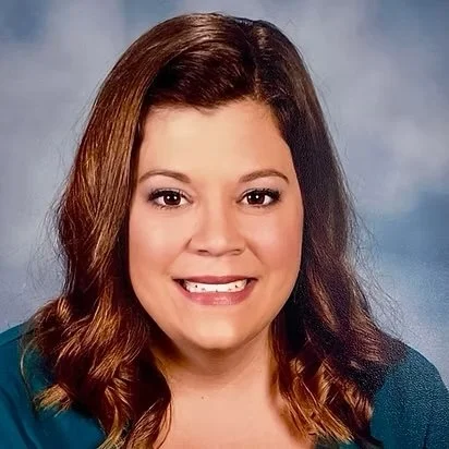A woman with shoulder-length brown hair, smiling, wearing a teal top, against a cloudy blue background.