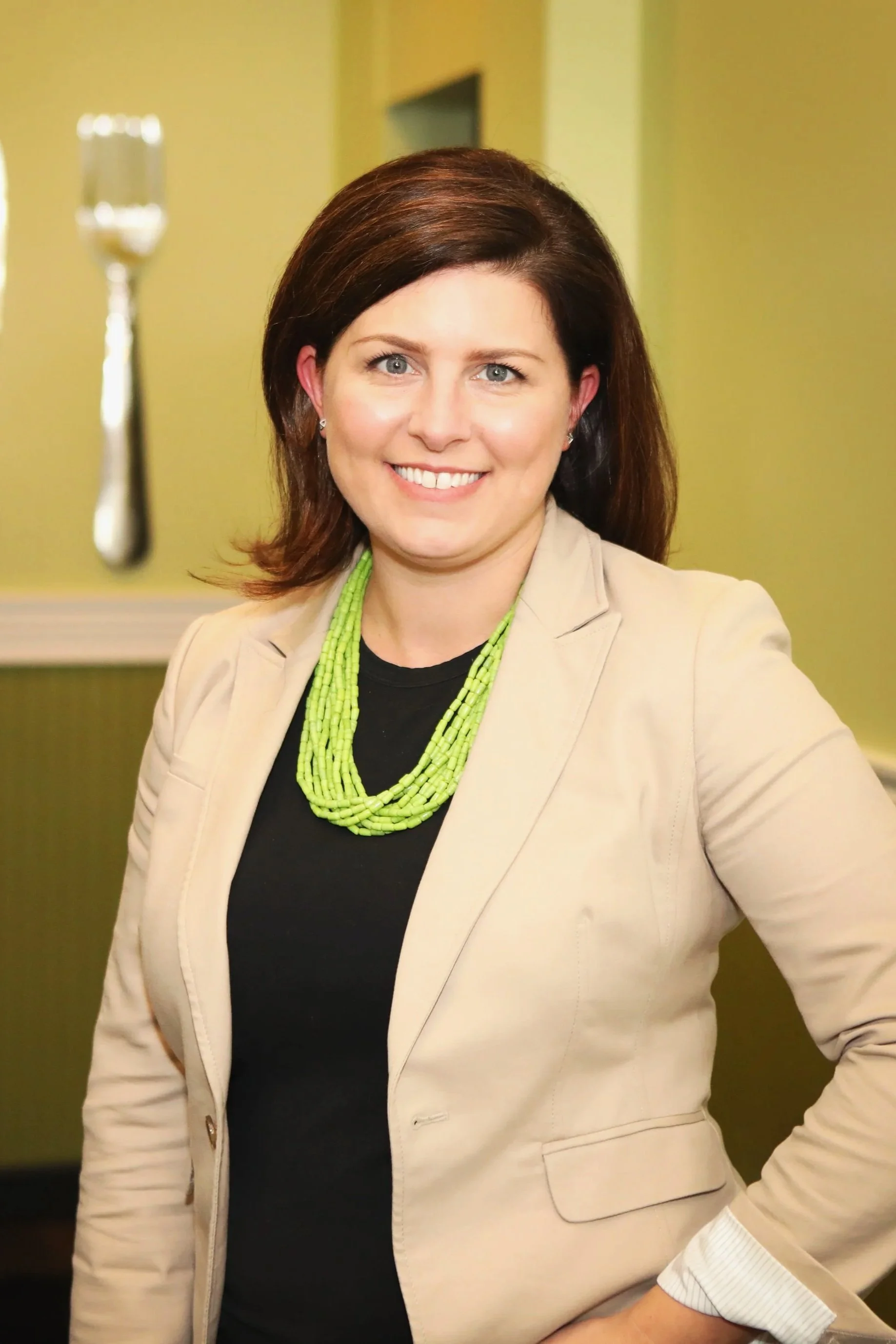 A woman with brown hair and blue eyes, smiling, wearing a beige blazer, black shirt, and a multi-strand green necklace, standing in a room with yellow walls and a large spoon decoration in the background.