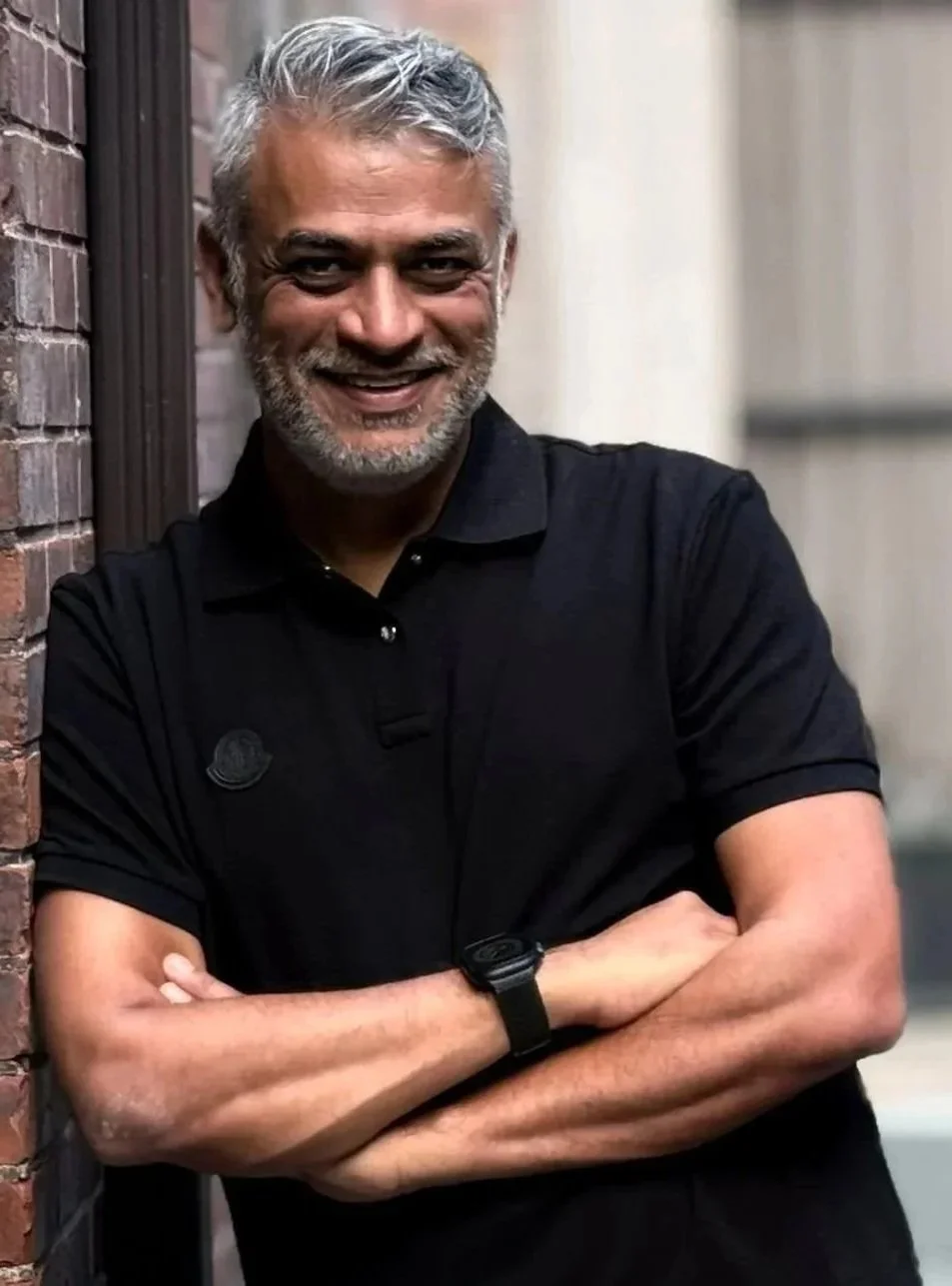 A smiling middle-aged man with gray hair and beard, wearing a black polo shirt, crossing his arms and leaning against a brick wall in an urban setting.