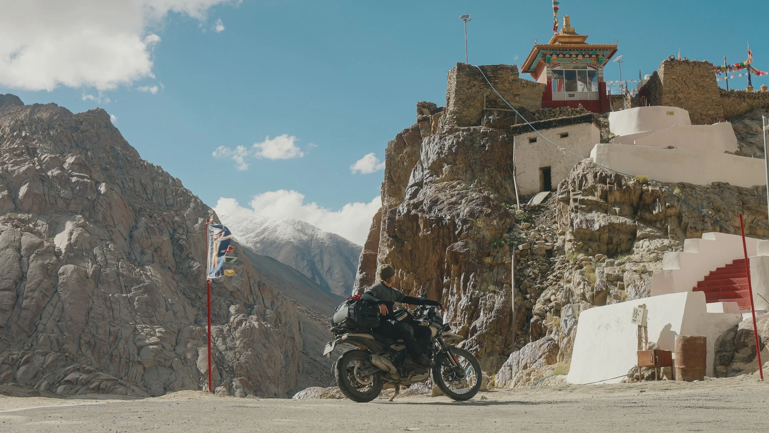 A motorcyclist riding a motorcycle with luggage in a mountainous area with rocky terrain, prayer flags, and a small building with colorful decorations on a cliff.
