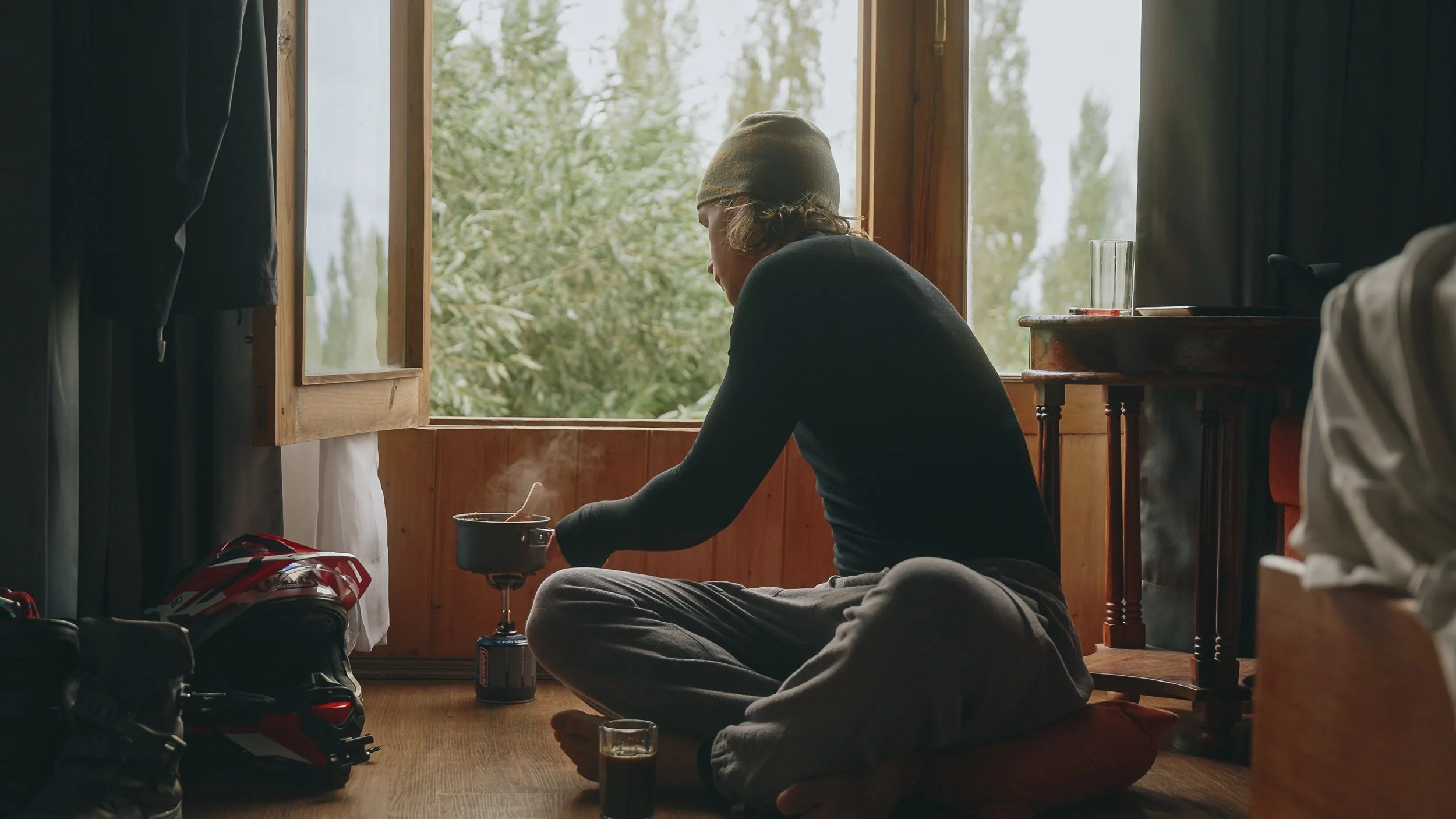 A person sitting cross-legged on a wooden floor by an open window, preparing camping or outdoor equipment, with a helmet and a drink nearby.