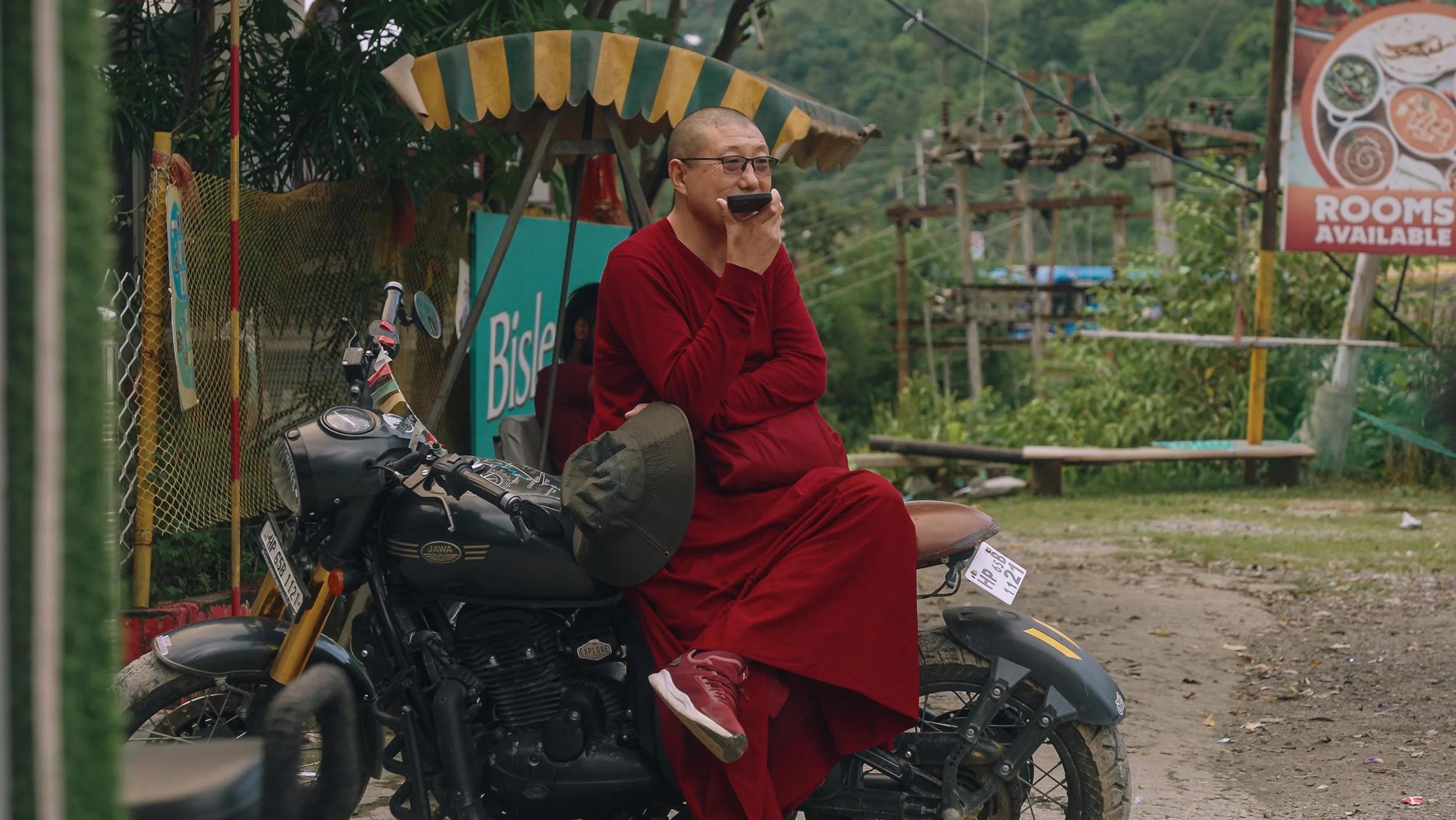 A person wearing a maroon robe and glasses sitting on a black motorcycle with a saddlebag, holding a phone near their mouth. The motorcycle is parked next to a fence with a yellow and green umbrella and some signs in the background.