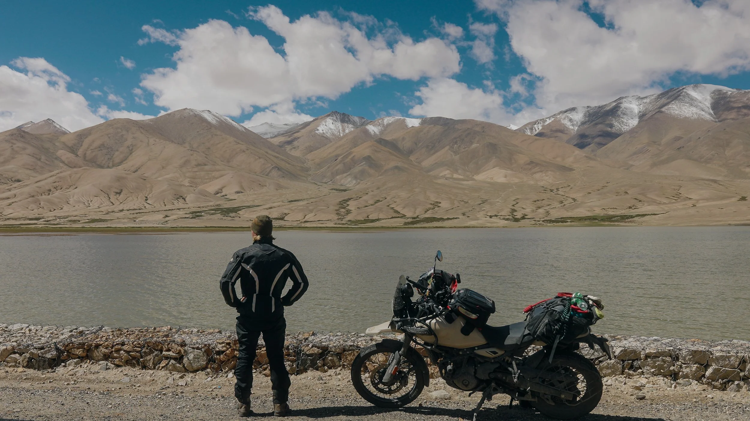 A person standing by a body of water with their back to the camera, facing a mountain range under a partly cloudy sky, with a motorcycle and luggage on the roadside.