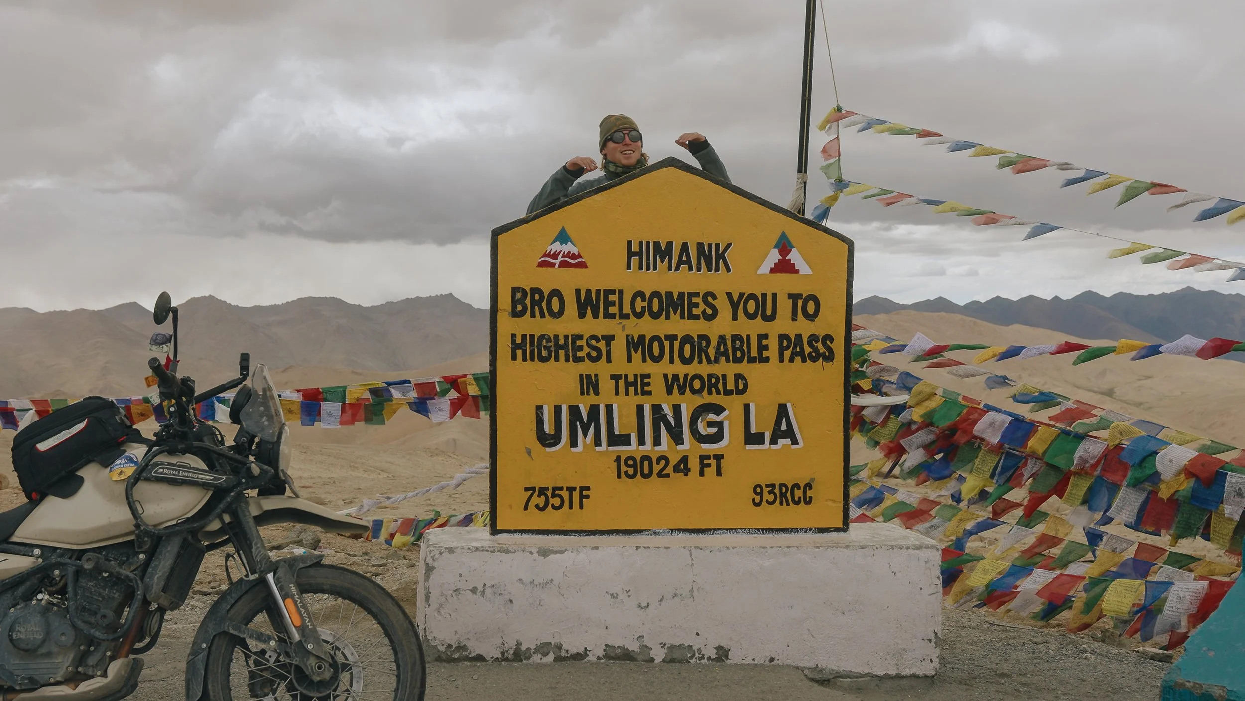A woman on a motorcycle standing behind a yellow sign at Umling La, the highest motorable pass in the world at 19,024 feet, with colorful prayer flags and mountainous terrain in the background.