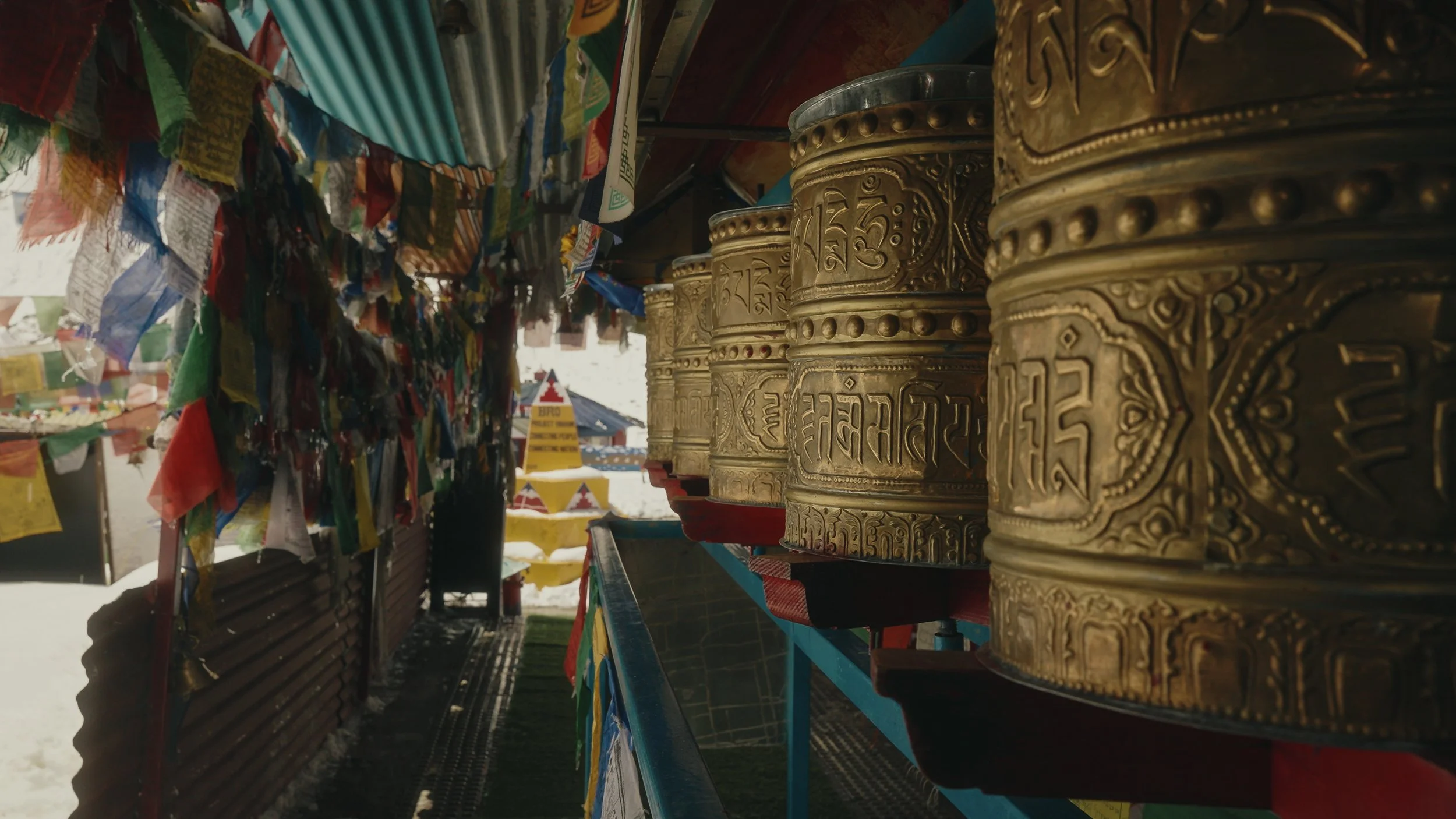 A row of golden prayer wheels with intricate carvings in a marketplace setting, surrounded by colorful prayer flags.