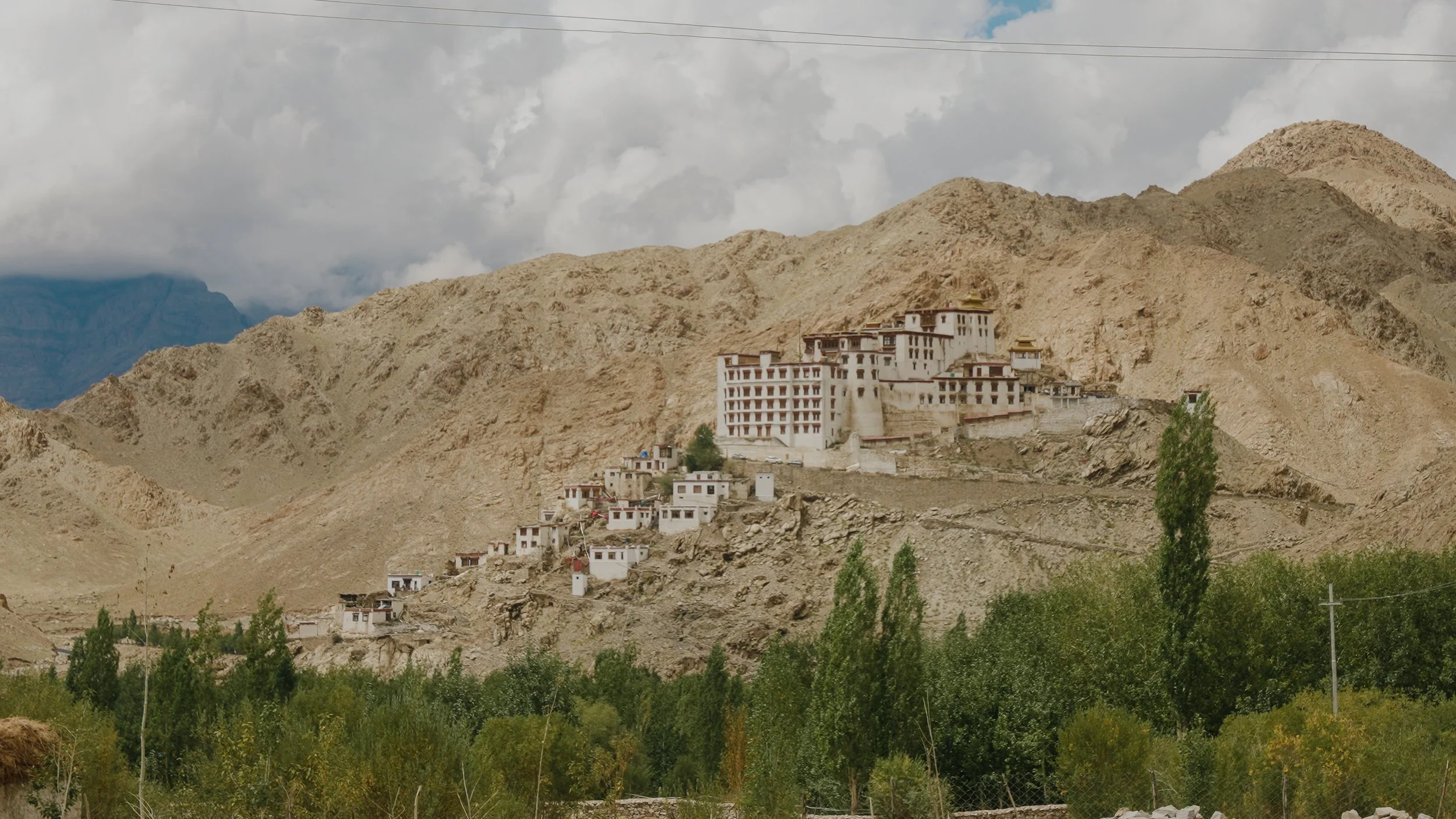 A mountainside landscape with a large white building complex, smaller structures, and some greenery in the foreground.