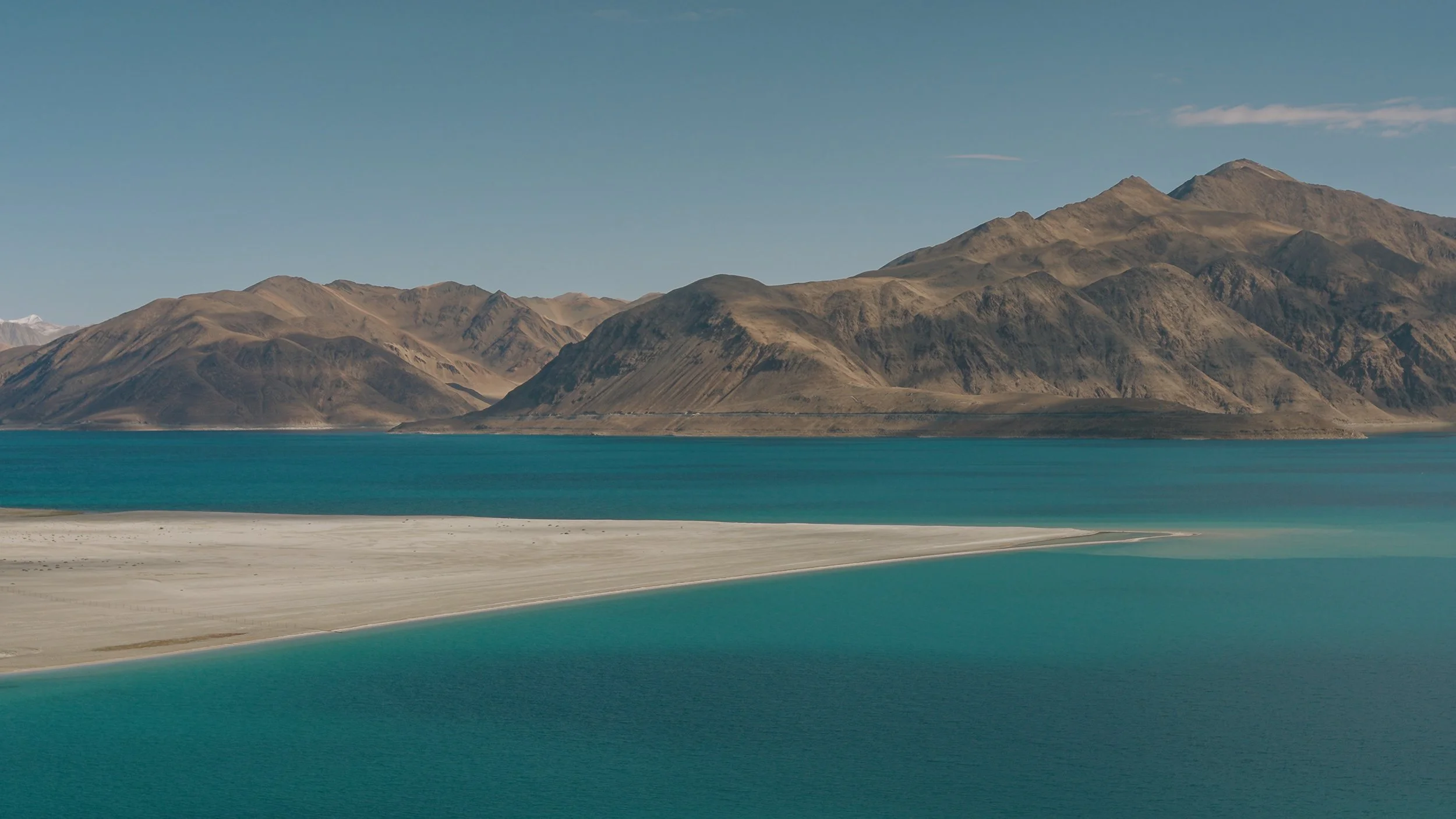 Scenic view of a mountain range with brown and gray peaks behind a large, turquoise lake with sandy shores, under a clear blue sky.