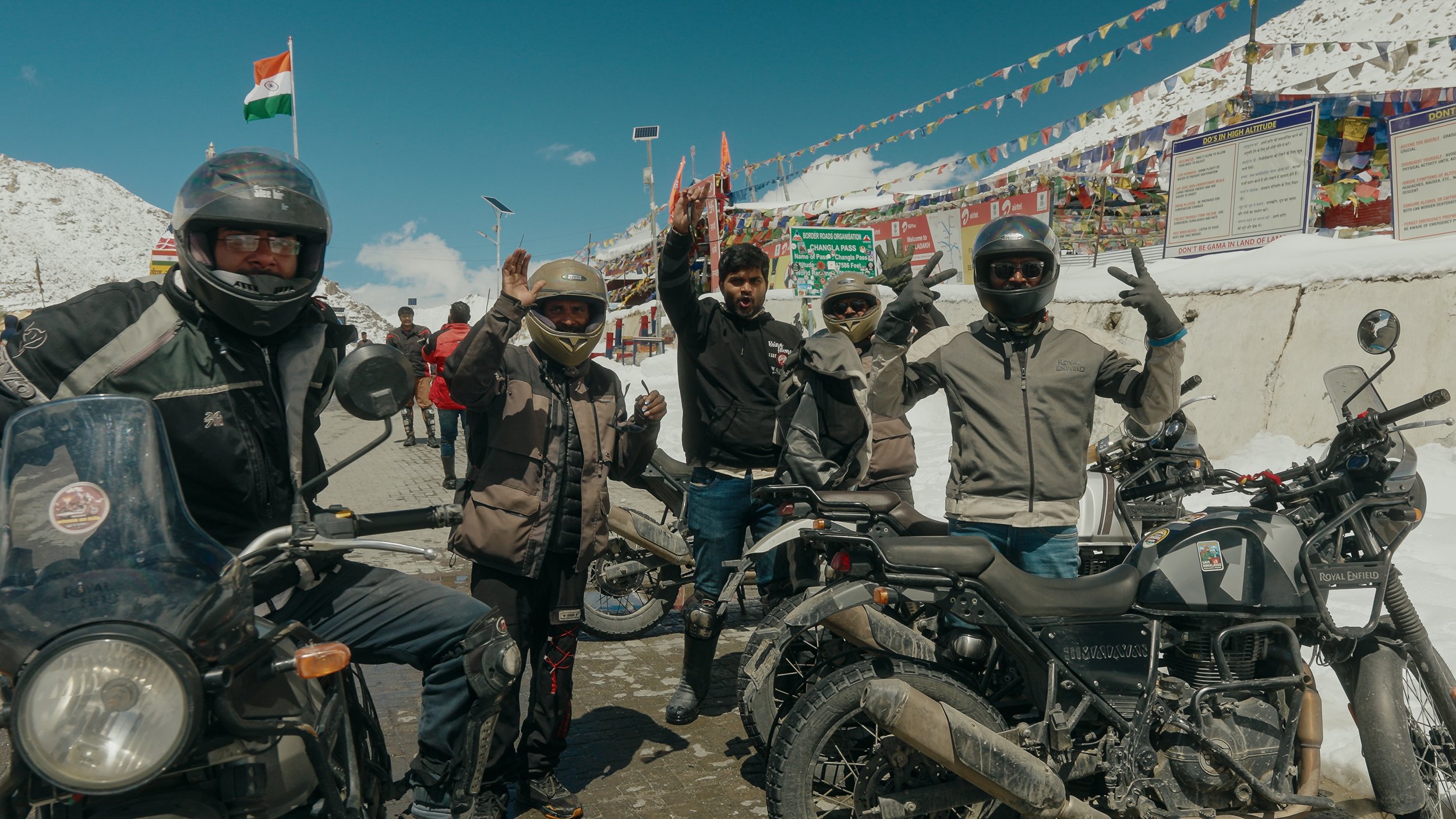 Group of motorcyclists wearing helmets and jackets, posing with peace signs at high altitude mountain location with colorful flags and signs in the background.