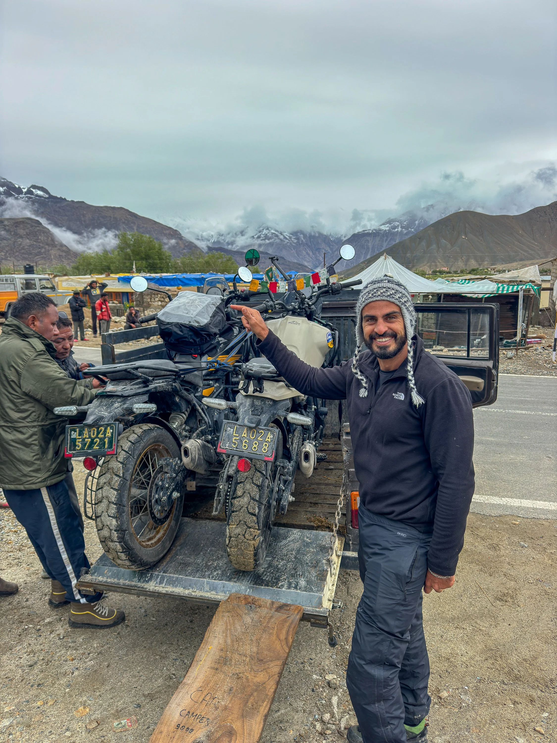 A man smiling and holding the handle of a motorcycle on a trailer, with two other men working on the motorcycle, in a mountainous area with tents and vehicles in the background.