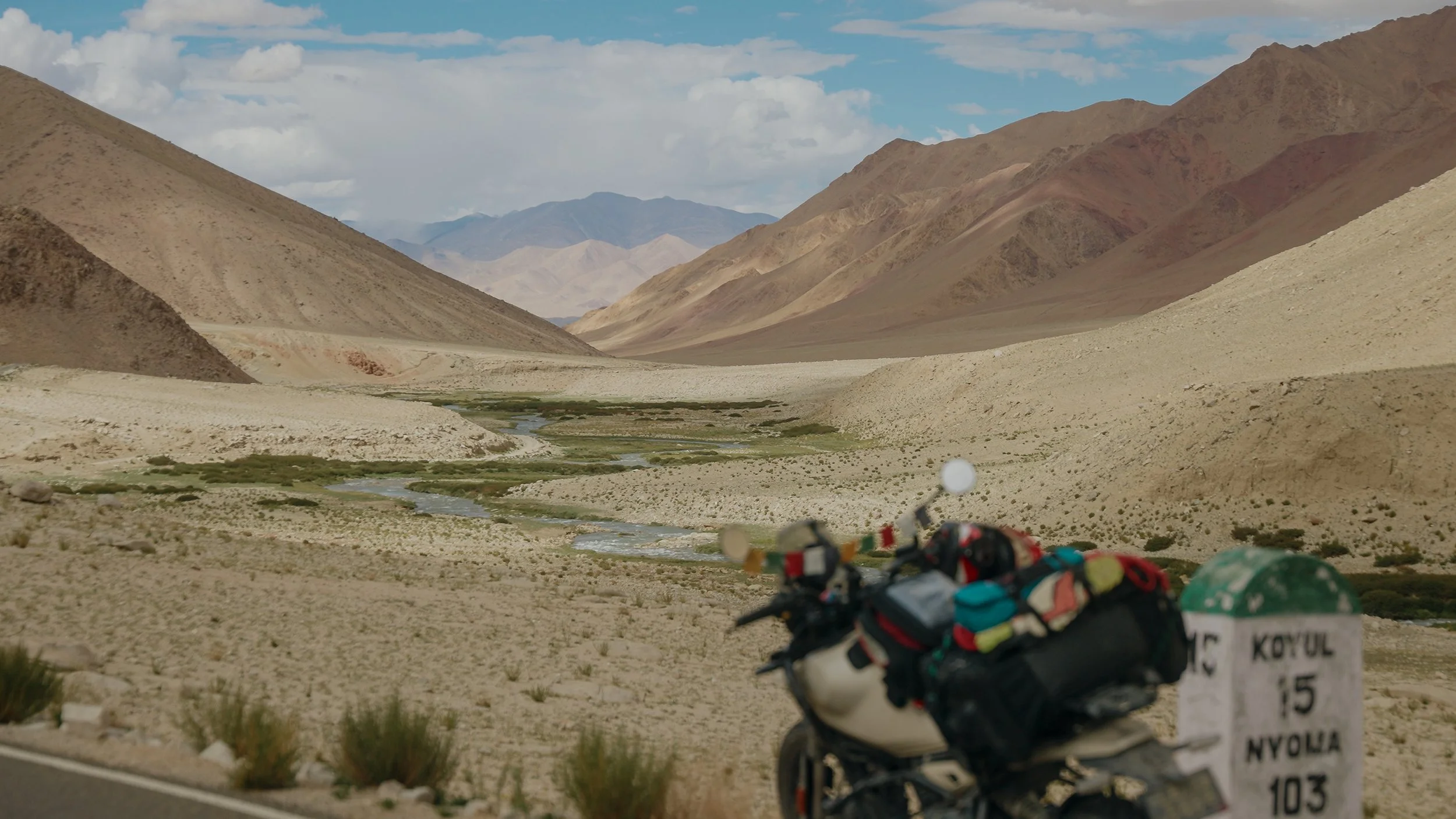 Motorcycle with colorful gear and personal items parked on the side of a desert mountain landscape with a winding river.