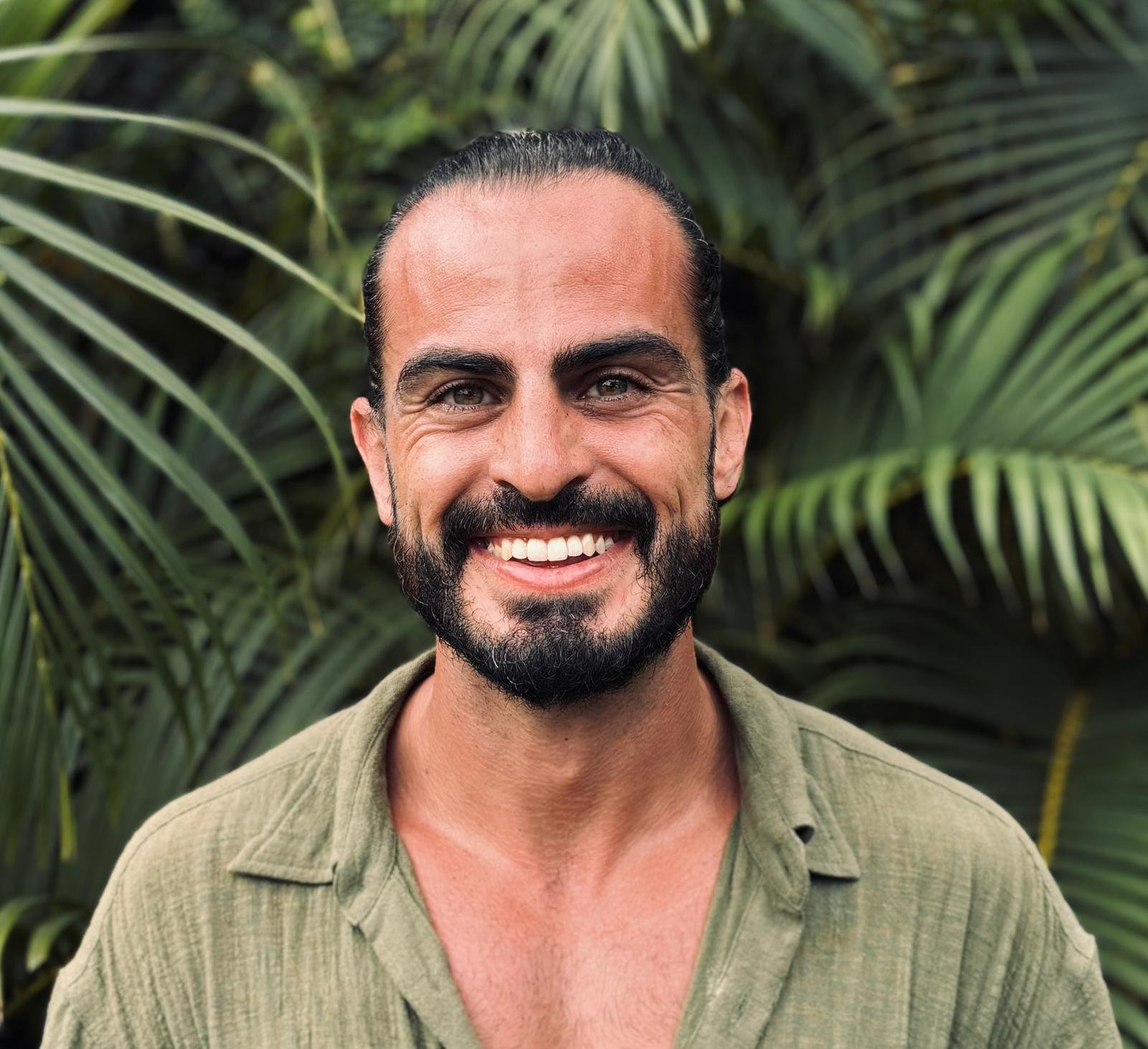 A smiling man with long dark hair slicked back, a beard, wearing a green shirt, stands outdoors in front of tropical plants.